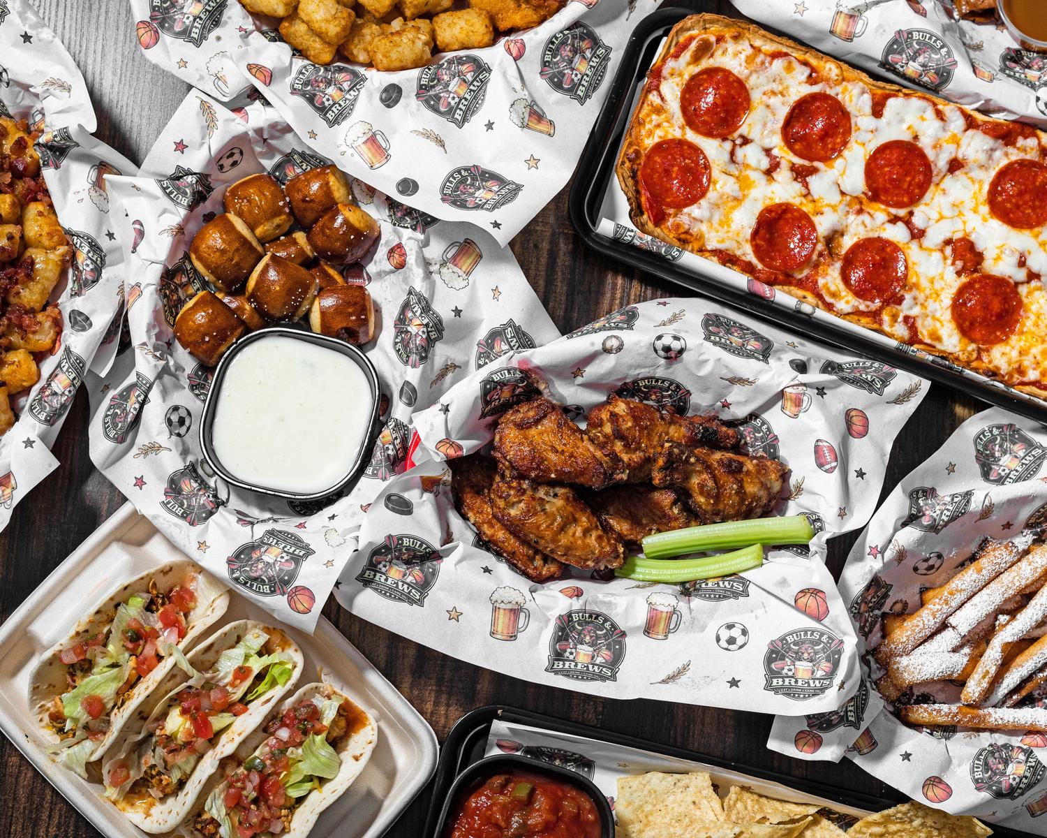 Overhead spread of Bulls & Brews wings, pepperoni flatbread, tacos, loaded tots, pretzel bites and funnel fries on a wooden table