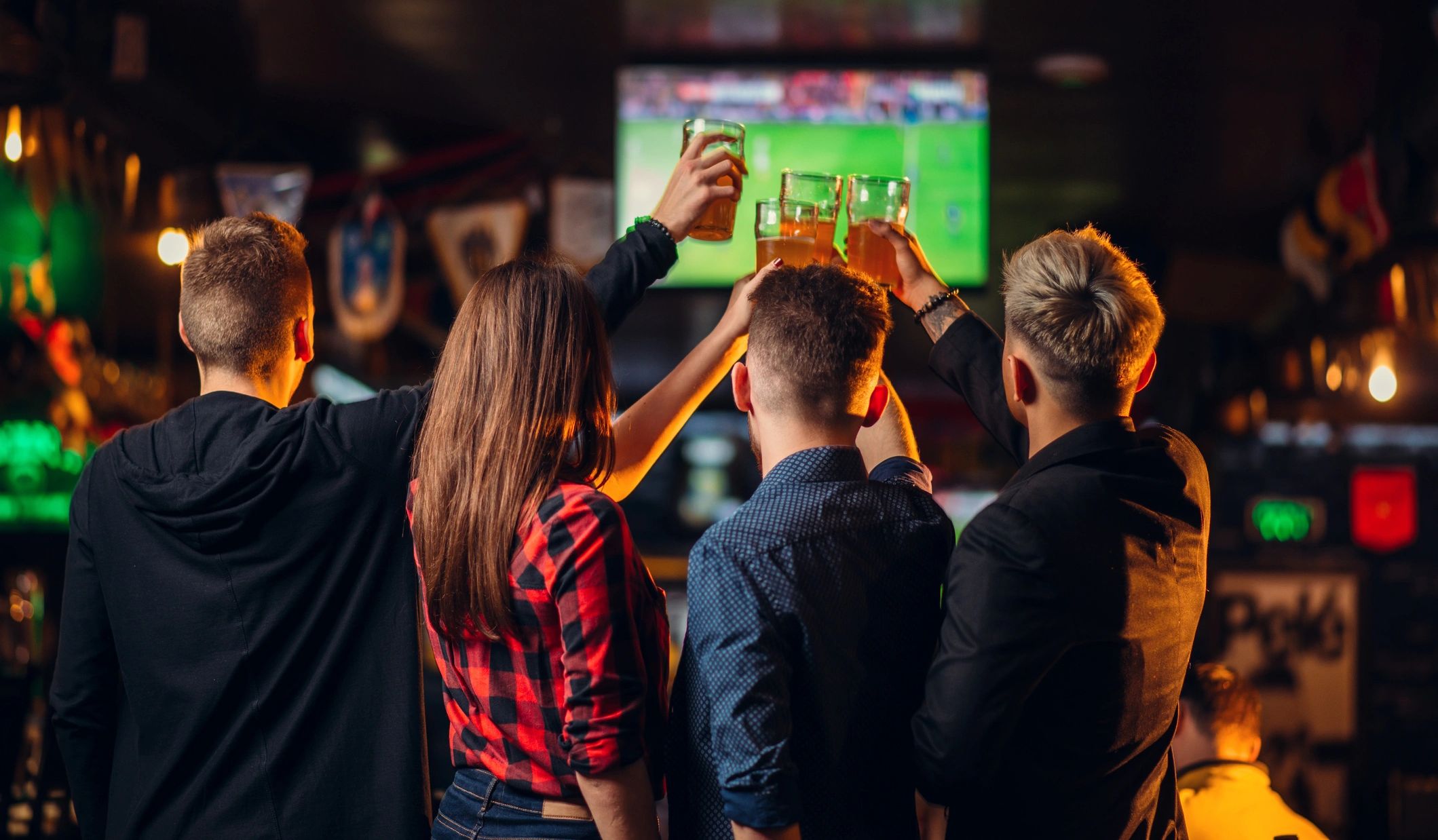 Four friends at the bar raising beer glasses in a toast in front of a game on TV